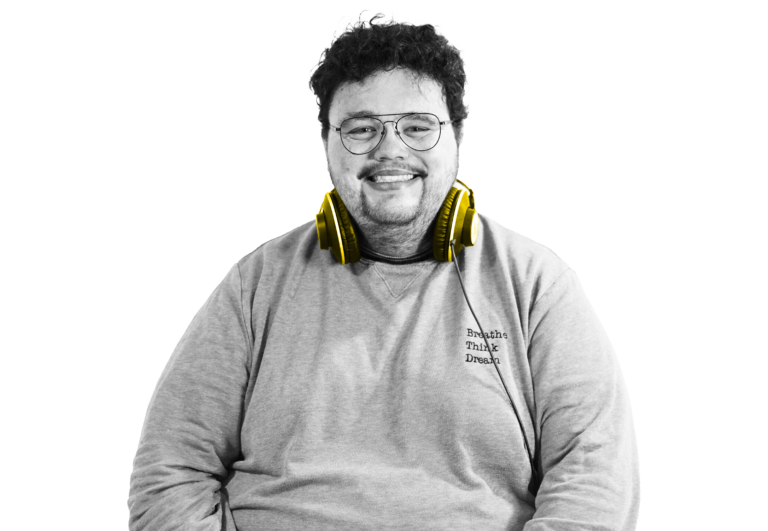Un homme souriant aux cheveux bouclés portant des lunettes, un sweat-shirt gris et des écouteurs jaunes autour du cou, isolé sur un fond blanc.